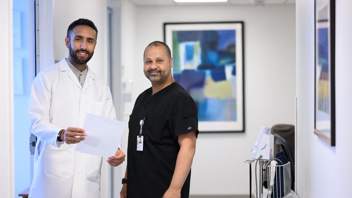 Two smiling healthcare workers stand in a bright clinical room, one holding a document and the other wearing black scrubs.