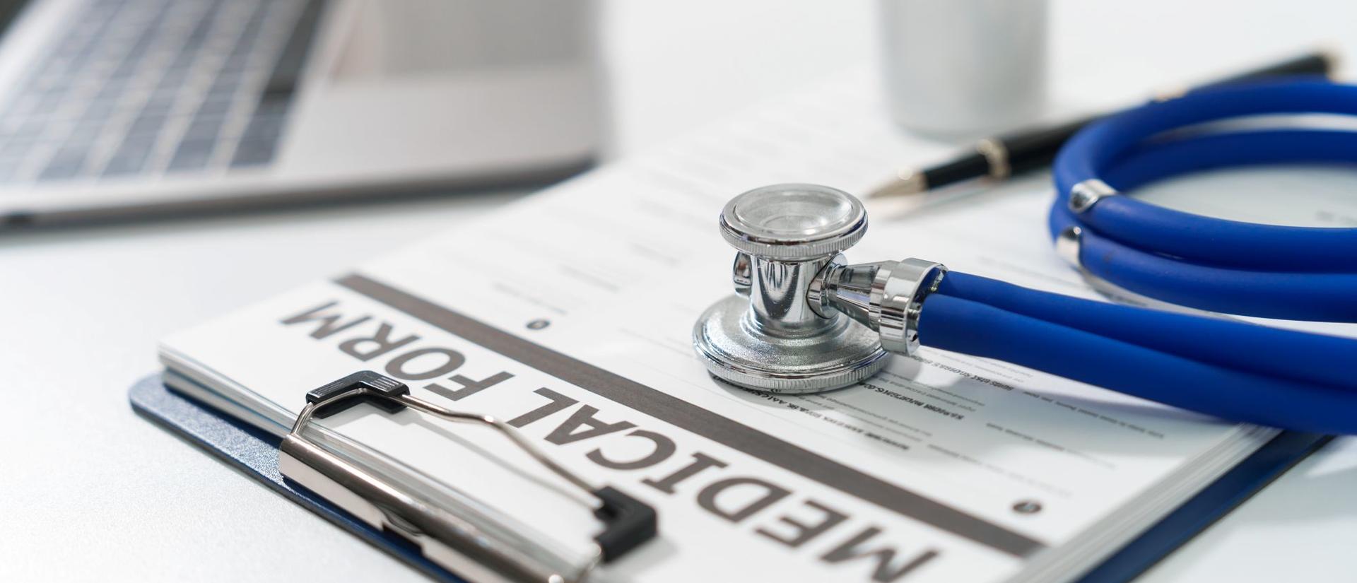 A stethoscope and a pen lie on a medical form on a clipboard, next to a laptop, on a white surface.