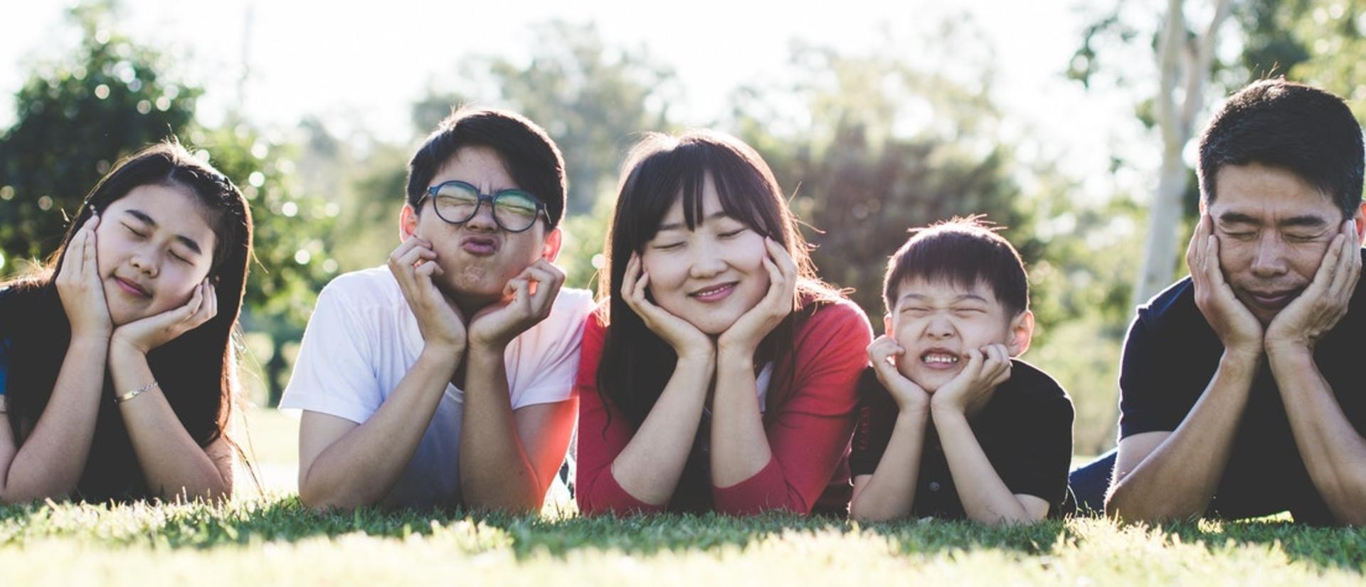 A family of five people laying on grass on their stomachs and their faces in their hands making being silly.